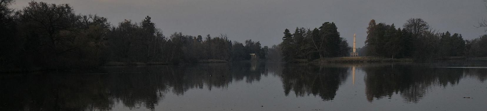 Blick über großen Teich auf Obelisk im Hintergrund in Gehölzumrandung. Landschaftspark Lednice.
